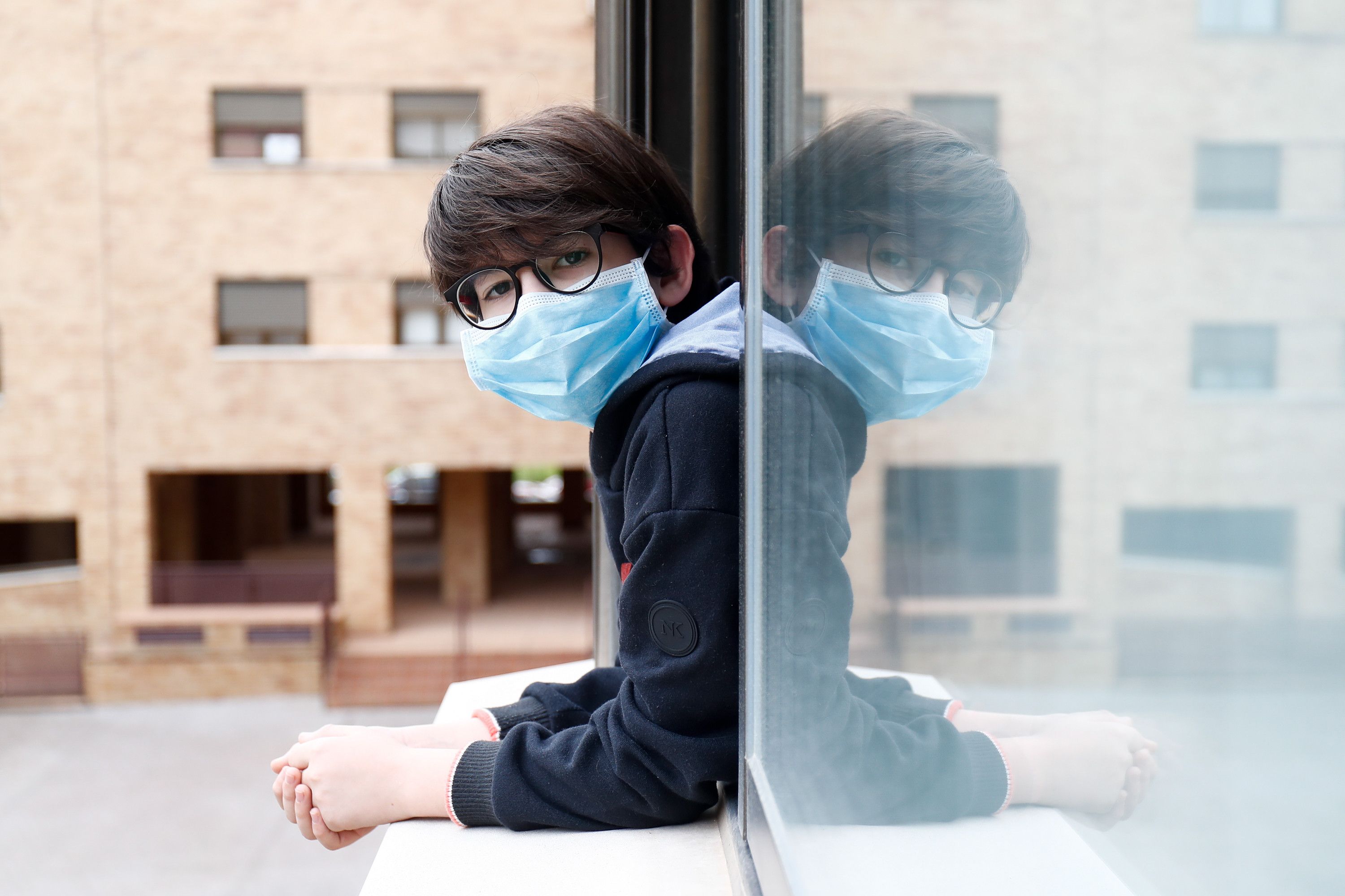 Un niño con una mascarilla se asoma a la ventana de su casa durante el confinamiento por el coronavirus. EP